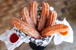 Churros goreng bertabur gula kayu manis disajikan dalam keranjang dengan saus celup karamel dan stroberi