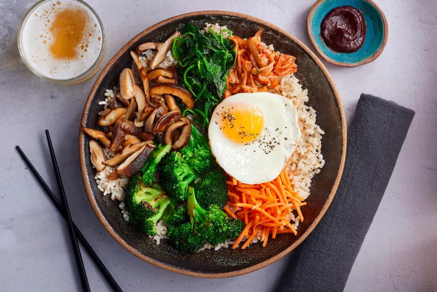 Semangkuk bibimbap Korea berisi nasi, brokoli, wortel, jamur shiitake, bayam, kimchi, dan telur mata sapi, disajikan dengan saus gochujang dan minuman di sampingnya